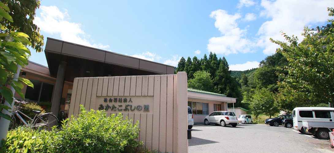 山林に囲まれた介護施設の建物外観と駐車場。木製の看板が見える。