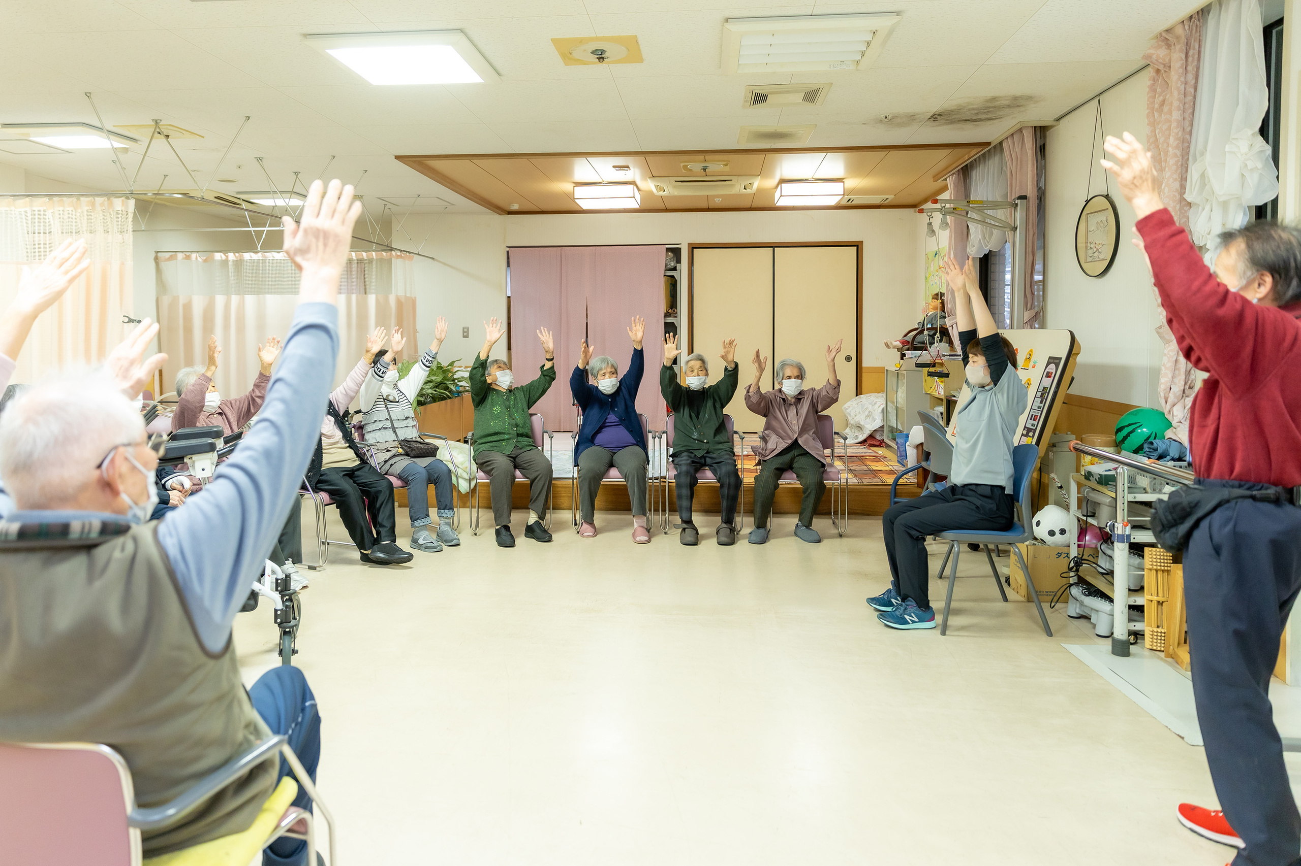 介護施設での体操レクリエーション。複数の入居者と職員が腕を上げる動作を行っている。