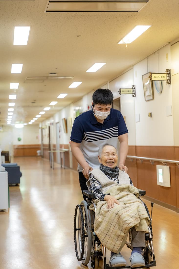 職員が高齢入居者を車椅子で廊下を移動介助している日常風景